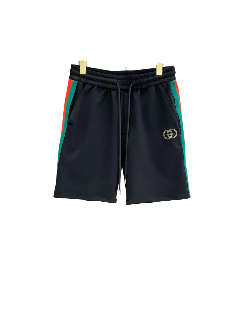 Black Gucci shorts with green and red side stripes and a gold interlocking G logo on the left leg, hanging on a clear hanger against a gray tiled wall.